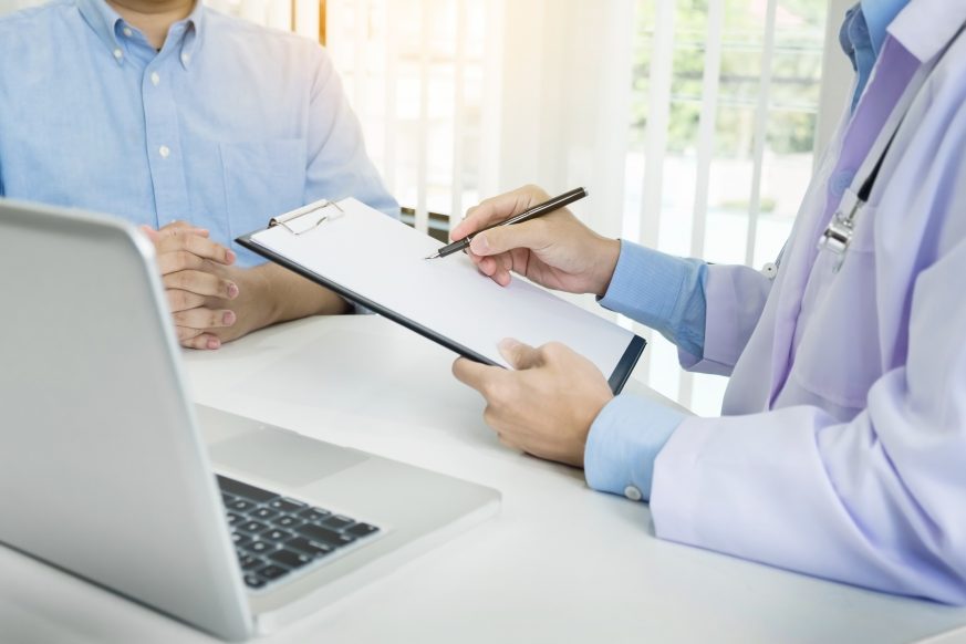 doctor talking to patient holding clipboard next to laptop